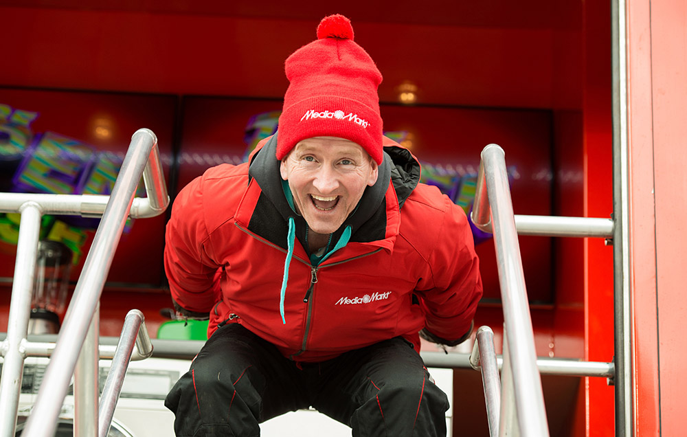 Speaker Eddie the Eagle in der Skirsprunghocke in roter Jacke und schwarzer Hose.