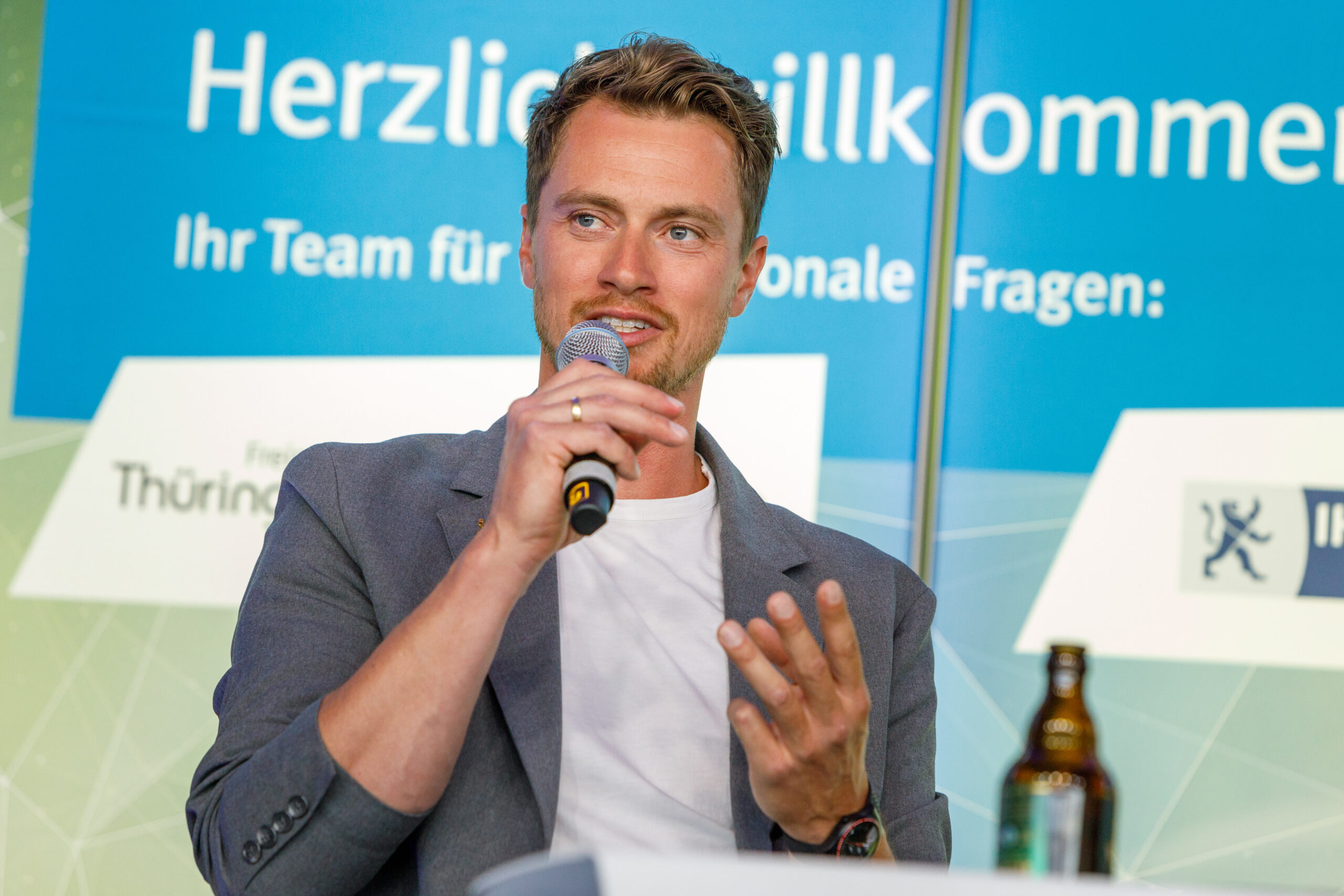 Dreifach-Olympiasieger Johannes Ludwig im grauen Sakko mit weißem T-Shirt und Mikro in der Hand.